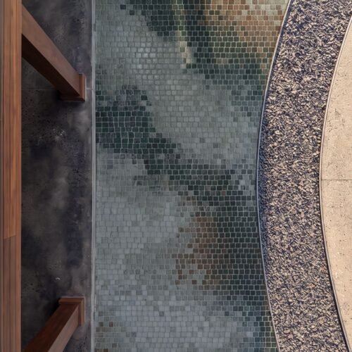 A close overhead view of an abstract stone walkway mosaic in green, taupe, and rust tesserae running alongside a curved gravel path.