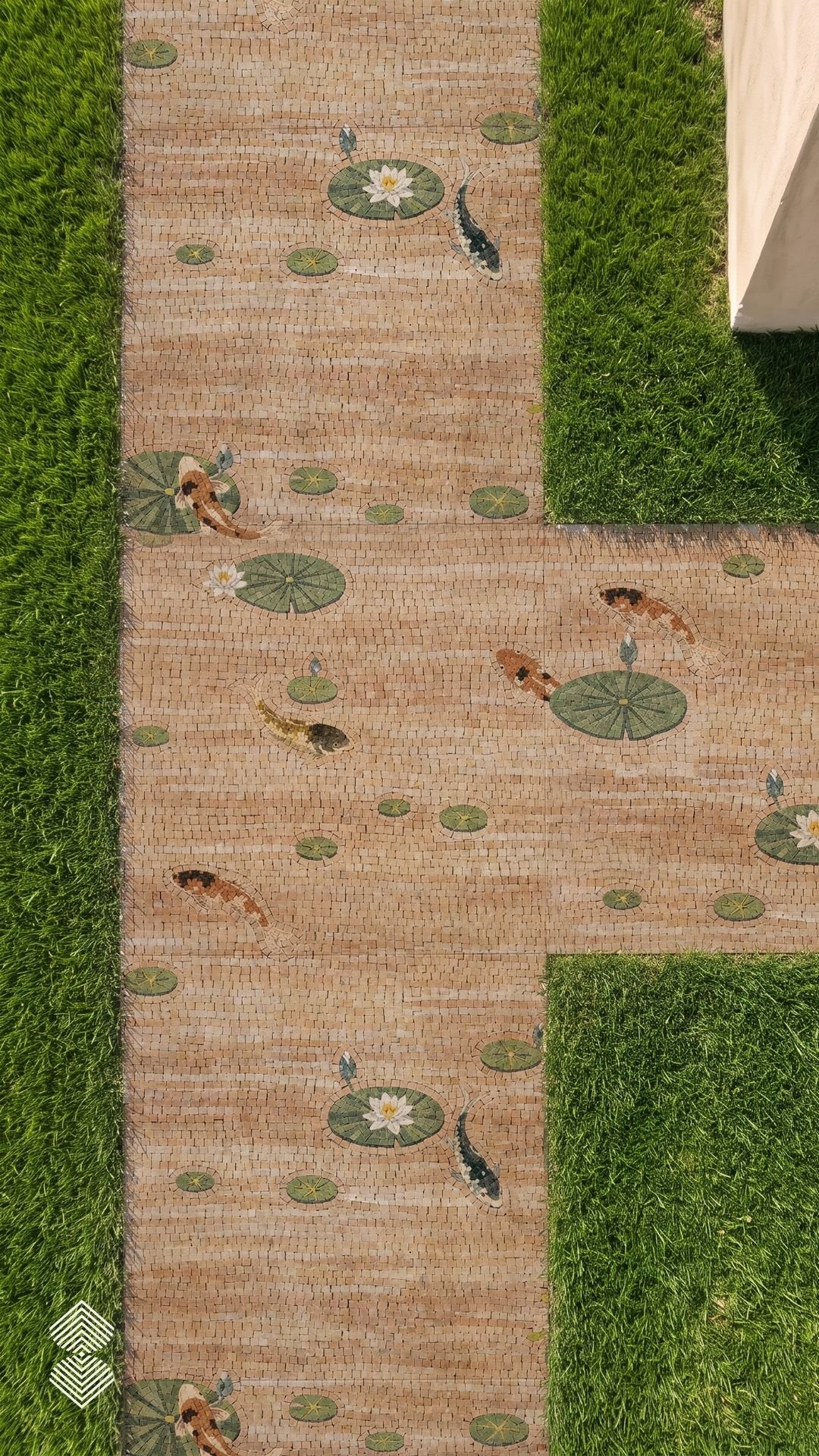 Overhead view of a tan stone garden path inlaid with mosaic koi fish, green lily pads, and white water lilies set across a green lawn.