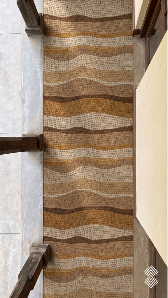 Top-down perspective of a mosaic walkway featuring flowing wave patterns in shades of cream, tan, and dark brown stone.