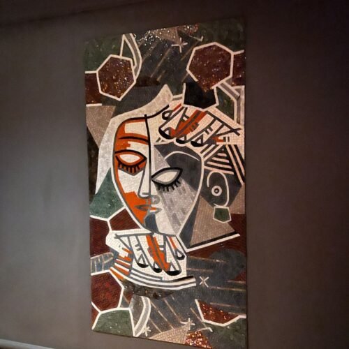 A cubist face mosaic mural in charcoal, rust and cream tones mounted on a dark cafe wall.