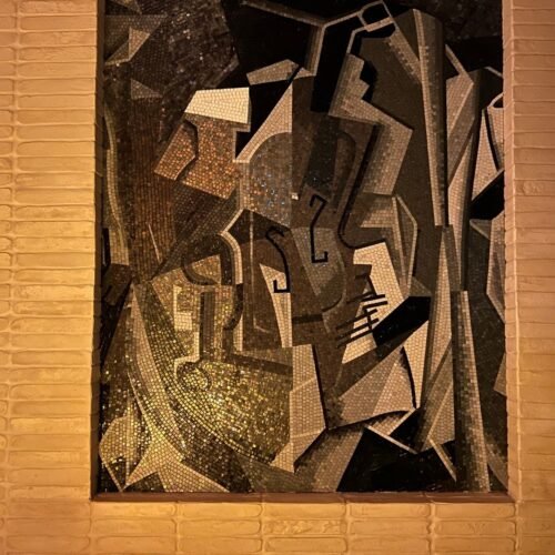 A cubist mosaic wall art panel of fragmented gray and gold glass tesserae set in a recessed niche.