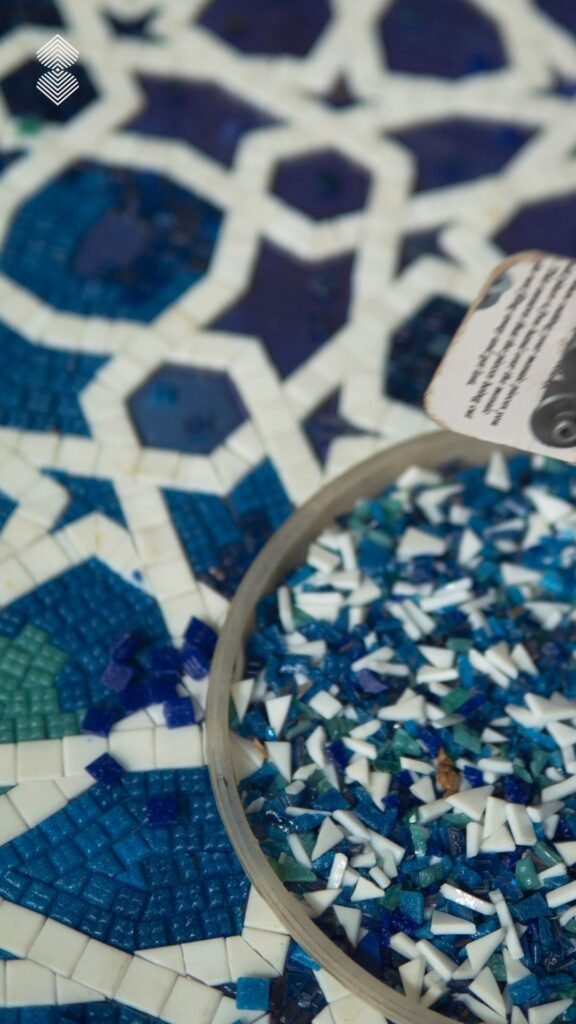 A close-up of a tray filled with small blue and white mosaic pieces resting on a partially completed geometric star mosaic pattern.