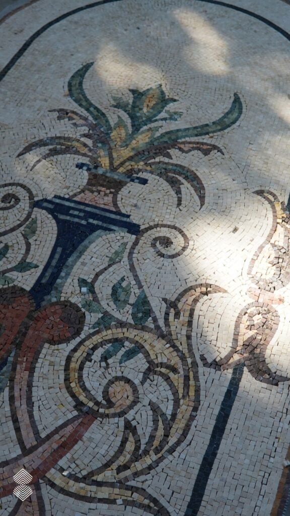 High-angle view of a large oval mosaic floor inlay depicting a decorative vase with leafy green plants and sweeping terracotta flourishes.