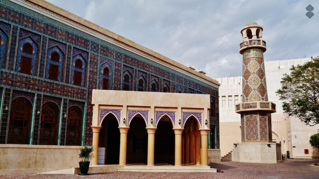 A grand mosque exterior heavily adorned with rich, dark blue patterned mosaic tiles, featuring arched walkways and a detailed minaret.
