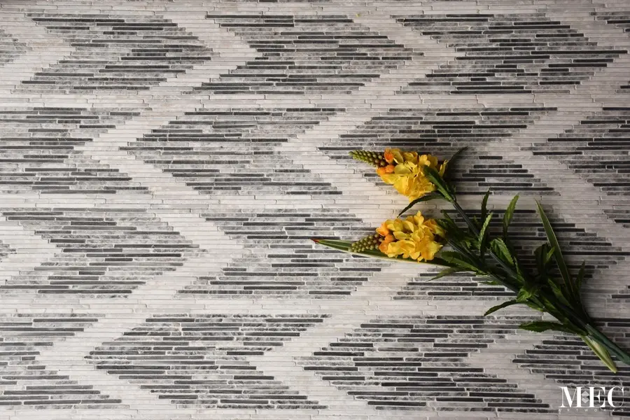 A flat lay view of horizontal thin mosaic tiles forming a grey and white chevron ikat pattern with yellow flowers resting on the surface.