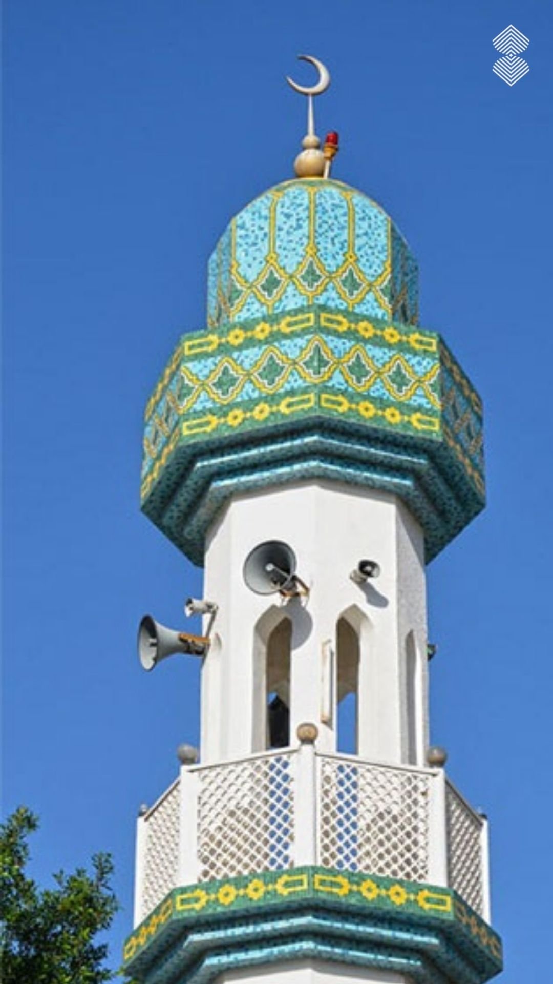 A white mosque minaret featuring a decorative top and balcony accented with light blue, green, and yellow geometric mosaic tiles.