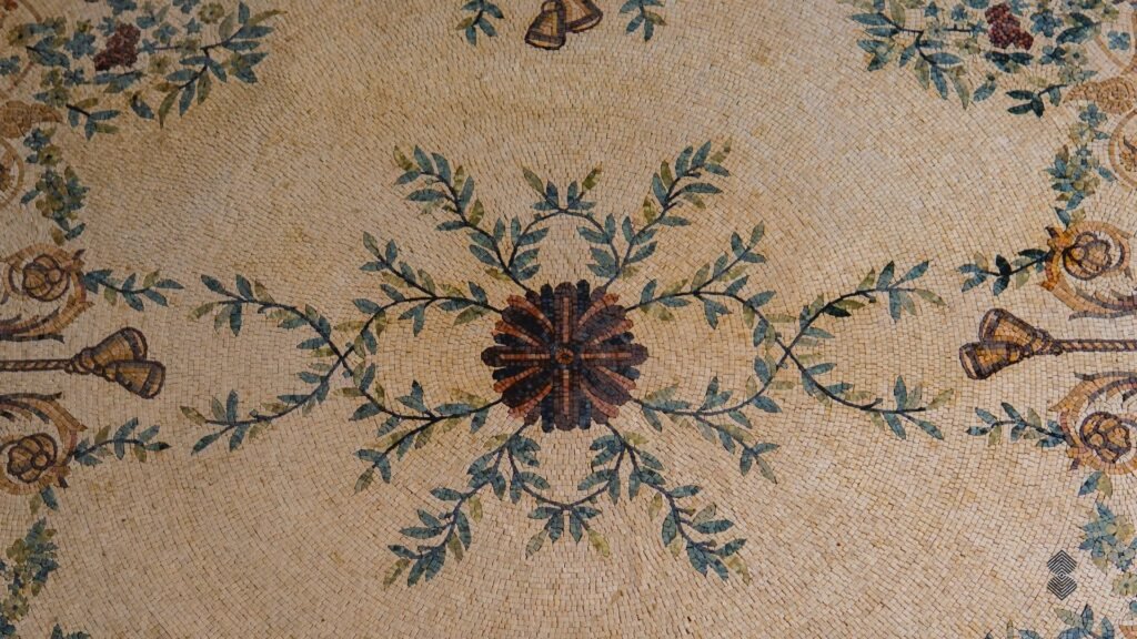 Overhead view of an ornate rectangular marble mosaic rug featuring a central floral emblem, green leafy branches, and decorative borders.