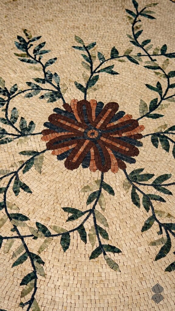 Close-up of a marble mosaic floor showing a dark orange and brown flower surrounded by leafy green vines on a beige background.
