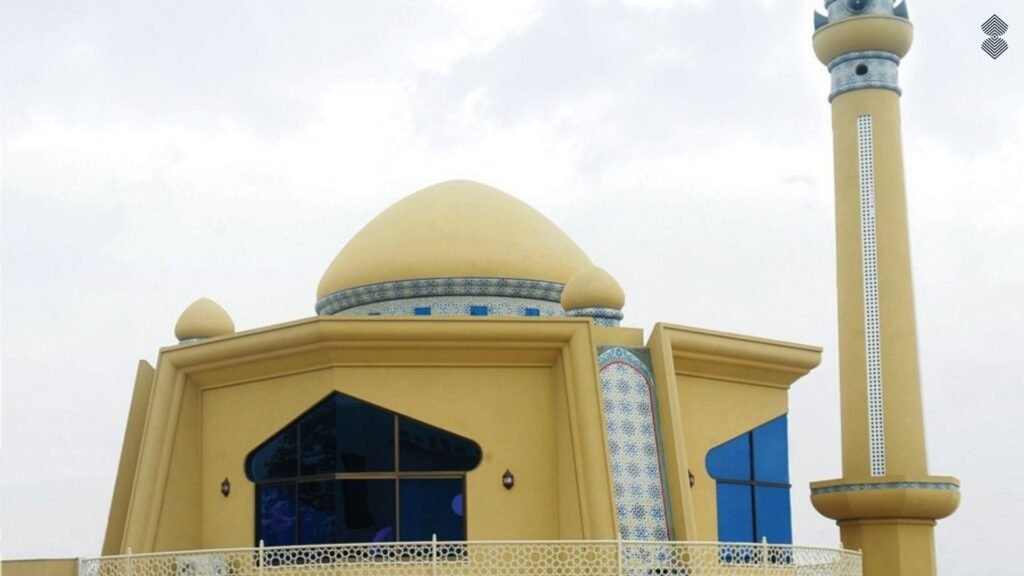 A pale. yellow modern mosque with a smooth dome and a tall minaret, accented with a thin band of blue and white mosaic tilework.