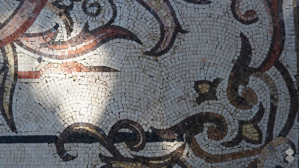 Close-up of a stone mosaic floor showcasing intricate dark brown and terracotta scrollwork curves against a off-white tile background.