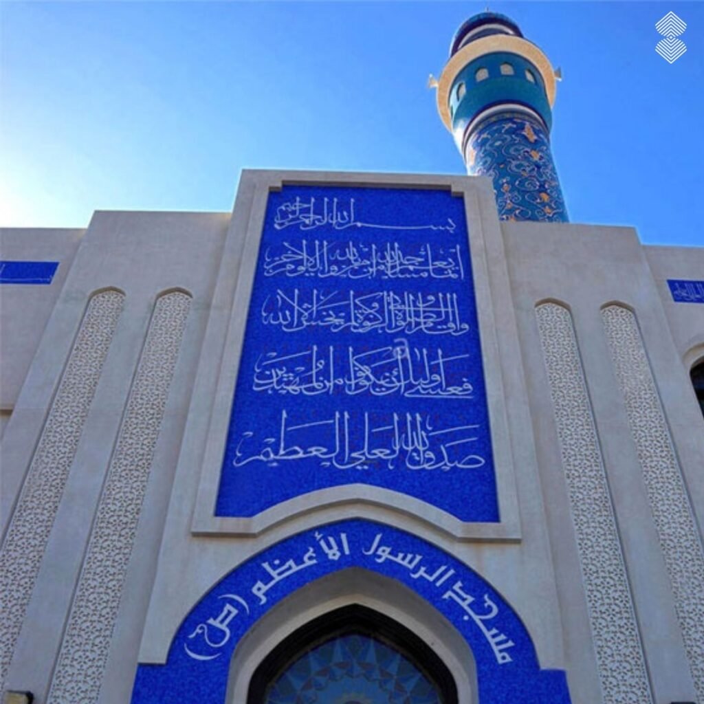 A prominent bright blue mosaic panel displaying white Arabic calligraphy on a light-colored mosque wall, with a decorated minaret in the background.
