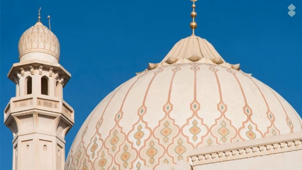 A smooth, light beige mosque dome decorated with subtle pastel pink and green geometric tracery, set against a bright blue sky.