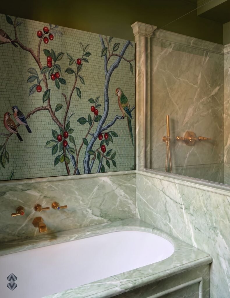 A green marble bathtub with brass fixtures beneath a mosaic wall mural featuring birds, branches, and red berries on a light green background.