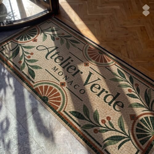 A tumbled stone art deco logo floor mosaic with script lettering, sunburst corners and olive branch motifs set at a doorway threshold.