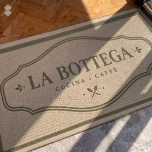 A cafe logo mosaic mat in tumbled cream and olive stone set into a doorway threshold.