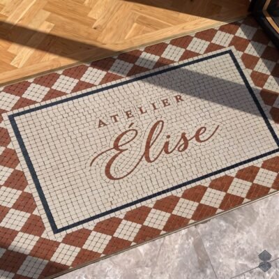 A script logo entryway mosaic in tumbled cream and terracotta stone, framed by a charcoal border and a diamond lattice.