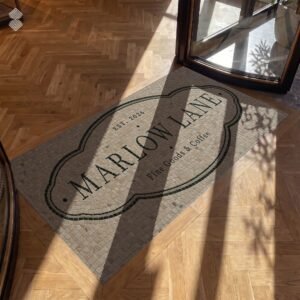 An oval crest entrance logo floor mosaic in tumbled beige stone set into a wooden entryway.
