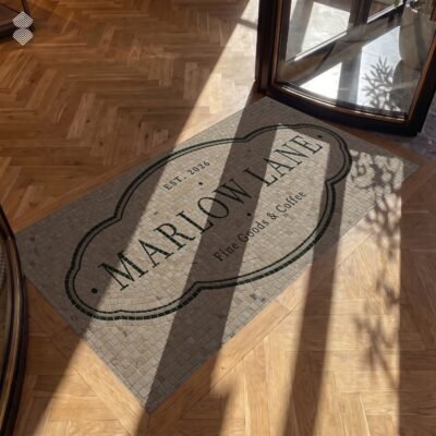 An oval crest entrance logo floor mosaic in tumbled beige stone set into a wooden entryway.